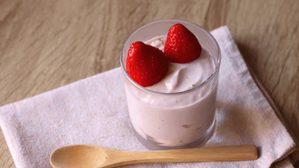 Mousse à la fraise onctueuse surmontée de deux fraises fraîches dans un verre transparent, avec une cuillère en bois reposant sur un tissu